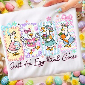May include: White t-shirt featuring four cartoon geese dressed for Easter. Each goose is in a decorative frame with floral and patterned backgrounds. The text "Just An Egg-cited Goose" is printed in a script font.