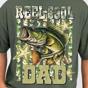 May include: Olive green t-shirt with a graphic of a largemouth bass and the words "REEL COOL DAD" in a camouflage design. The fish is green and yellow with an open mouth. Stars are in the background.