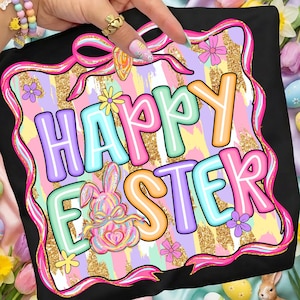 以下が含まれることがあります： カラフルな「Happy Easter」グラフィックがプリントされた黒いTシャツ。パステルカラーの文字、ウサギ、花、金色のグリッターの背景が特徴で、ピンクのリボンで縁取られています。お祝いのイースターをテーマにしたアパレルアイテムです。