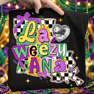 Könnte beinhalten: Schwarzes T-Shirt mit einem farbenfrohen Grafikdesign. Das Design zeigt die Worte "La Weez Ana" in verschiedenen Farben und Schriftarten, zusammen mit einem Discokugelherz, Schleifen und einer Fleur-de-Lis. Das Design ist im Stil einer Mardi Gras-Feier.