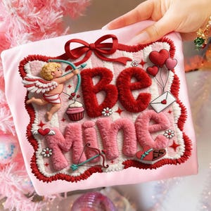 Peut inclure: Un coussin carr&eacute; rose avec les mots "Be Mine" dans une police de caract&egrave;res moelleuse et textur&eacute;e. Le motif comprend un n&oelig;ud rouge, un cupidon avec un arc et des fl&egrave;ches, des c&oelig;urs et un cupcake. La bordure est rouge et blanche.