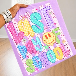 May include: A rectangular, lavender-colored blanket with the words "LAST DAY OF SCHOOL" in colorful, bubble letters. The design includes a smiley face, stars, and flowers. The blanket has a striped pattern.