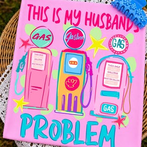 Op de afbeelding: Een roze canvas met de tekst "THIS IS MY HUSBAND'S PROBLEM" en illustraties van benzinestations. Het kunstwerk bevat het woord "GAS" in cirkels en sterren. Een blauwe haarclip met bloemdetails bevindt zich rechtsboven.