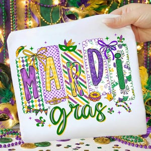 May include: White fabric featuring a vibrant "Mardi Gras" design. The text is in purple, yellow, and green, with decorative elements such as masks, beads, and a trumpet. The design is held by a hand.