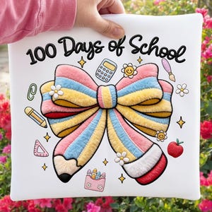 Könnte beinhalten: Weißes Kissen mit dem Text "100 Days of School" und einem großen, farbenfrohen Schleifendesign. Die Schleife besteht aus rosa, gelben und blauen Streifen. Weitere Illustrationen zum Thema Schule sind ein Bleistift, ein Taschenrechner und ein Apfel.