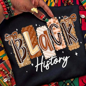 May include: Black t-shirt featuring the words "BLACK History" in a decorative font. The letters are in various patterns and colours, including brown, beige, and plaid. The shirt is displayed on a colourful patterned fabric.