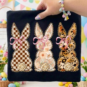 Puede incluir: Camiseta negra con tres diseños de conejos apilados. Cada conejo tiene un patrón diferente: a cuadros, floral y estampado de leopardo. Lazos rosas y detalles florales adornan el gráfico temático de Pascua.