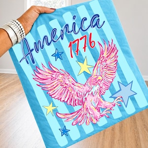 以下が含まれることがあります： 縦縞模様の薄い青色のブランケット。筆記体で「America」の文字と赤い「1776」の数字。ピンクと黄色のワシのグラフィックが中央に配置され、青と黄色の星がアクセント。愛国的なテーマ。