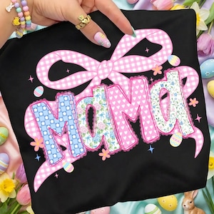 Op de afbeelding: Zwart T-shirt met een roze ruitjesstrik en het woord "Mama" in een decoratief lettertype. De letters zijn versierd met bloemen- en patroonontwerpen. Paaseieren en bloemen zijn verspreid rond het ontwerp.