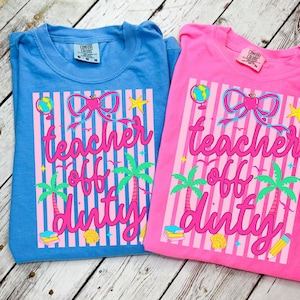 Op de afbeelding: Twee Comfort Colors t-shirts, één blauw en één roze, met de tekst "teacher off duty". Het ontwerp bevat een strik, palmbomen en een wereldbol. De shirts zijn opgevouwen op een licht houten oppervlak.