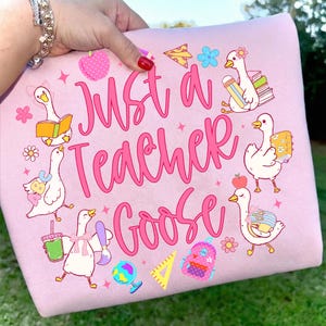 以下が含まれることがあります： ピンク色のスウェットシャツ。ピンク色の筆記体で「Just a Teacher Goose」と書かれています。デザインは、本、鉛筆、リンゴなどの学用品を持った漫画のガチョウが特徴です。スウェットシャツは人が持っています。