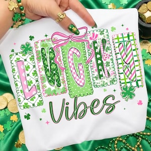 Pode incluir: Camiseta branca com as palavras "LUCKY VIBES" em verde. A palavra "LUCKY" está em blocos rosa e verde com padrões de trevo e florais. Um laço rosa está acima da letra "C". A camiseta é decorada com trevos.