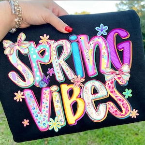 May include: Black sweatshirt with the words "SPRING VIBES" in colorful, whimsical lettering. The design includes floral accents, bows, and a butterfly, creating a cheerful and vibrant aesthetic. The letters have various patterns and textures.