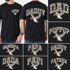 Op de afbeelding: Zwarte t-shirts met diverse tekstontwerpen, waaronder "DADDY", "DAD", "POPPPOP", "GRANDPA", "PAPA", "DADA", "PAPI" en "FATHER". Elk ontwerp bevat een eendenillustratie met uitgespreide vleugels. De tekst is in een camouflagepatroon.