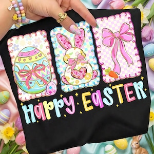 May include: Black t-shirt with a colorful "Happy Easter" graphic. The design features three framed images: an Easter egg, a bunny with a bow, and a pink bow with a carrot. The images have a pastel color scheme.