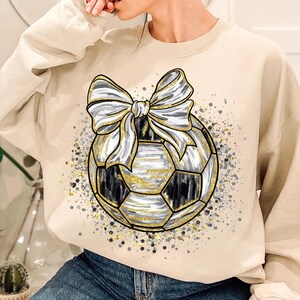 K&ouml;nnte beinhalten: Beiges Sweatshirt mit einem Fu&szlig;ball-Design und einer gro&szlig;en Schleife. Der Fu&szlig;ball ist schwarz, wei&szlig; und goldfarben, mit einem goldenen Umriss. Die Schleife ist wei&szlig; und grau. Das Design ist von goldenen und grauen Sprenkeln umgeben.