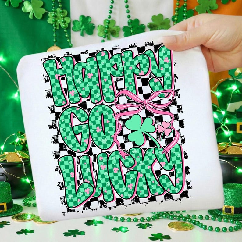 May include: White St. Patrick's Day themed shirt with the words "Happy Go Lucky" in green checkered letters with pink accents. A pink bow and shamrocks are also featured in the design. The shirt is held up by a hand.