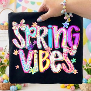 May include: Black t-shirt with the words "SPRING VIBES" in colorful, floral-themed lettering. The design includes butterflies, bows, and flowers in pastel colors. The shirt is held up by a hand.