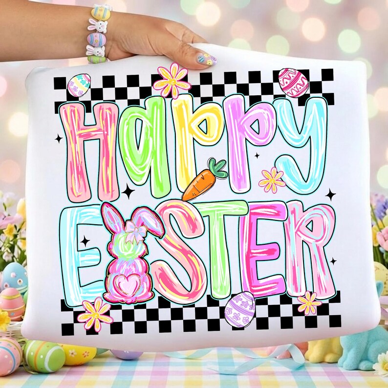 Może przedstawiać: Biała poduszka z napisem "Happy Easter" w kolorowych literach. Projekt zawiera kr&oacute;lika, marchewkę i pisanki, z czarno-białą szachownicą.