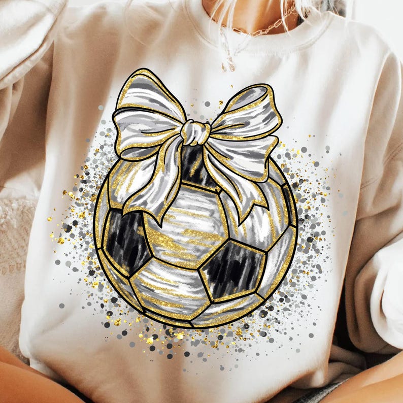 K&ouml;nnte beinhalten: Ein cremefarbenes Sweatshirt mit einem Fu&szlig;ball-Design und einer gro&szlig;en Schleife. Der Fu&szlig;ball ist schwarz und wei&szlig; mit goldenen Akzenten, und die Schleife ist silber mit goldenen Details. Das Design ist von goldenem und silbernem Glitzer umgeben.