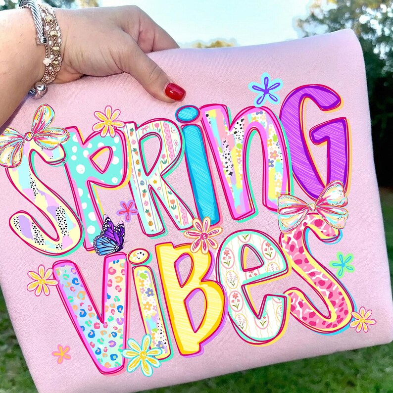 May include: A light pink sweatshirt with the words "SPRING VIBES" in colorful, patterned letters. The design includes butterflies, flowers, and bows. The sweatshirt is held up by a hand with a red painted nail.