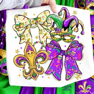 May include: White graphic tee featuring Mardi Gras themed designs. The design includes a gold fleur-de-lis, purple and green bows, a jester hat, and a mask. The design is embellished with gold glitter accents and star shapes.
