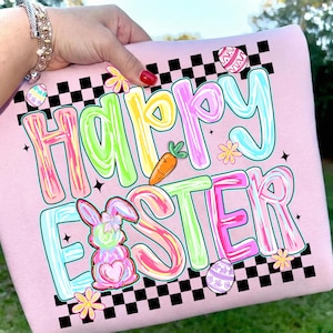 May include: A light pink sweatshirt with a colorful "Happy Easter" graphic. The design includes a bunny, carrot, and Easter eggs, with a black and white checkered pattern. The text is in a bubble letter style with pastel colors.