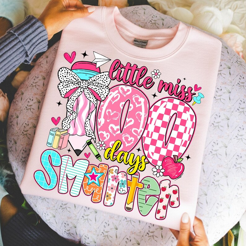 May include: A light pink sweatshirt with a colorful graphic design. The design includes the text "little miss 100 days smarter" with hearts, stars, and school-themed illustrations. The sweatshirt is held by a person.