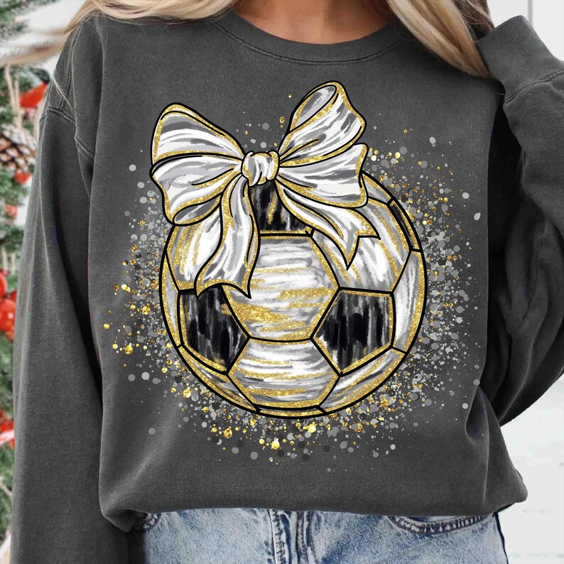 K&ouml;nnte beinhalten: Dunkelgraues Sweatshirt mit einem Fu&szlig;ball-Design und einer gro&szlig;en wei&szlig;en und goldenen Schleife. Der Fu&szlig;ball hat schwarz-wei&szlig;e Paneele mit goldenen Akzenten. Das Design ist von goldenem und silbernem Glitzer umgeben.