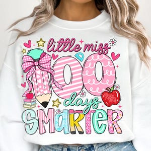 Puede incluir: Sudadera blanca con un gr&aacute;fico colorido que celebra los 100 d&iacute;as de escuela. El dise&ntilde;o incluye un l&aacute;piz grande con un lazo rosa, las palabras "little miss 100 days smarter", corazones, estrellas y una manzana.