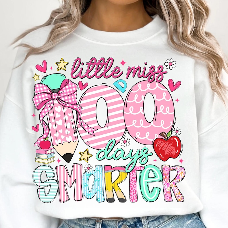 Puede incluir: Sudadera blanca con un gr&aacute;fico colorido que celebra los 100 d&iacute;as de escuela. El dise&ntilde;o incluye un l&aacute;piz grande con un lazo rosa, las palabras "little miss 100 days smarter", corazones, estrellas y una manzana.