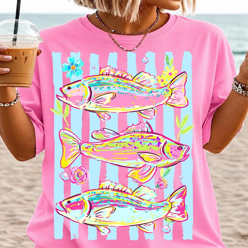May include: Pink t-shirt featuring a colorful illustration of three fish with floral accents and blue vertical stripes. The fish are in shades of pink, yellow, and blue. The shirt is a casual, relaxed fit.