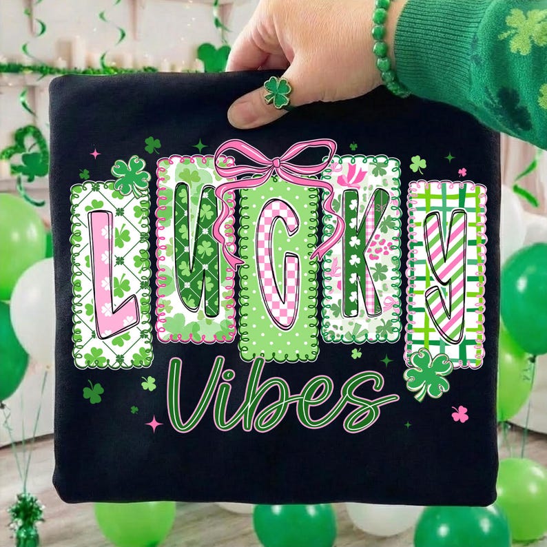 Op de afbeelding: Zwart T-shirt met de woorden "LUCKY Vibes" in groen en roze. De letters zijn versierd met klavers en patronen. Een roze strik staat boven de letter "C". Het shirt wordt door een persoon omhoog gehouden.
