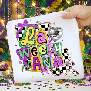 May include: White t-shirt with the text "La Weezu Ana" in colorful, block letters. The design includes a disco ball heart, a purple bow, and a checkered pattern, evoking a festive theme.