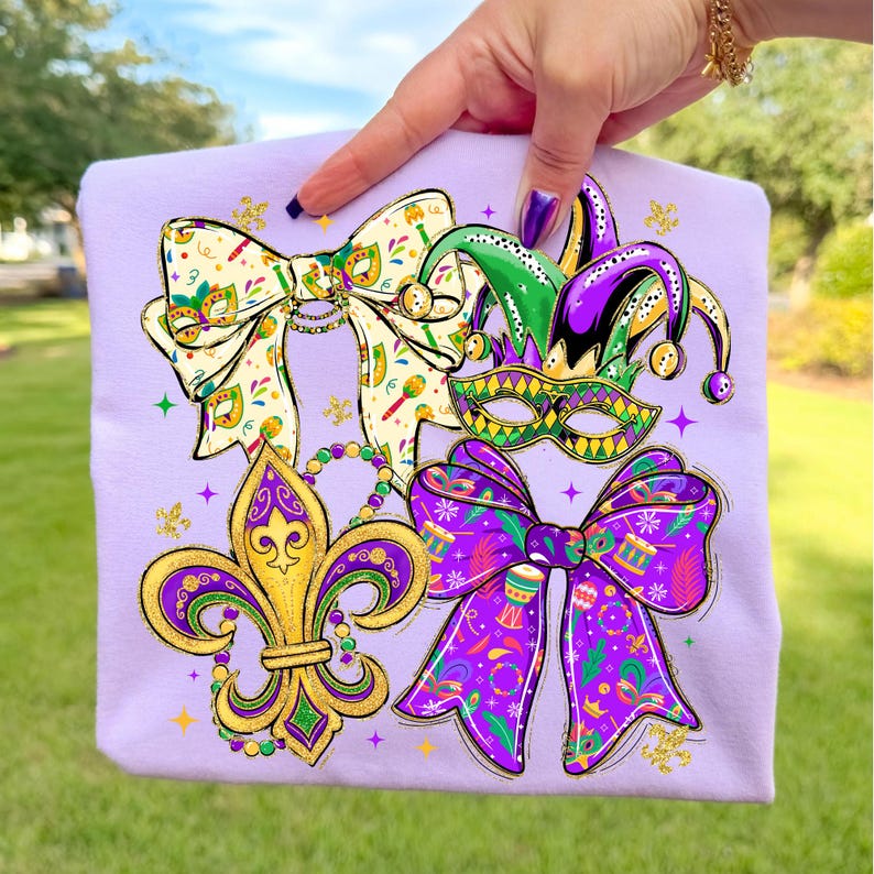 May include: A light purple sweatshirt featuring colorful Mardi Gras-themed graphics. The design includes a fleur-de-lis, bow ties, a jester hat, and a mask. The graphics are in gold, green, purple, and white.