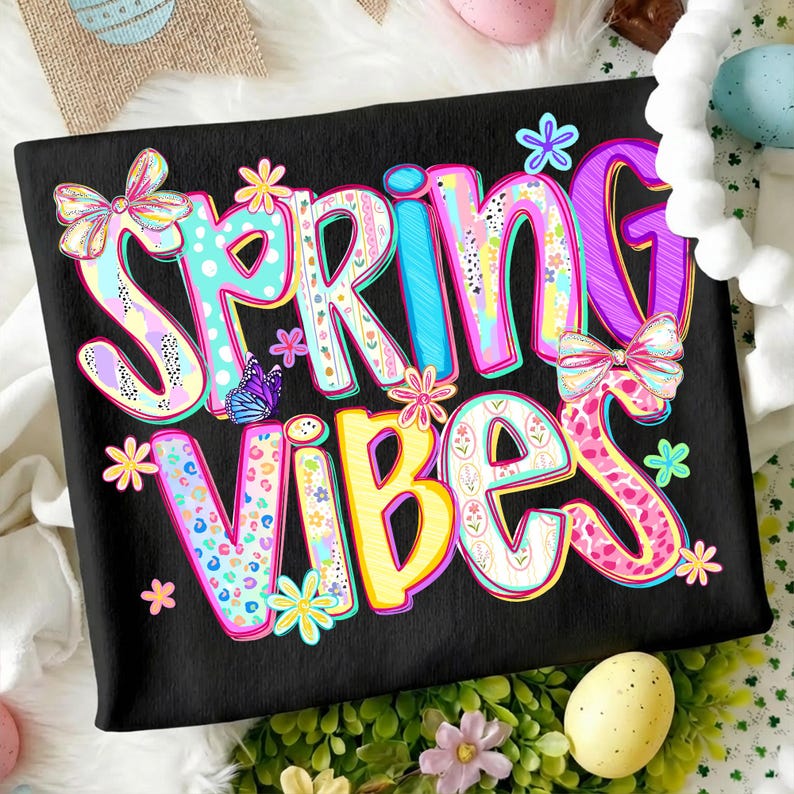 May include: Black t-shirt with the words "SPRING VIBES" in colorful, whimsical lettering. The design includes floral accents, bows, and a butterfly, evoking a cheerful, springtime theme.