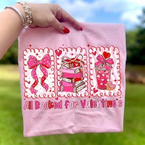 May include: Pink t-shirt with a Valentine's Day theme. The shirt features three framed illustrations: a bow, a stack of books, and a drink, all with hearts. The text "All Booked for Valentine's" is printed at the bottom.