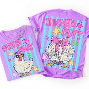 May include: Two purple t-shirts with chicken-themed graphics. One shirt says "GUESS WHAT" with a cartoon chicken wearing sunglasses. The other shirt says "CHICKEN BUTT" with a chicken's rear end and a pink bow. Both shirts have blue and pink stripes.