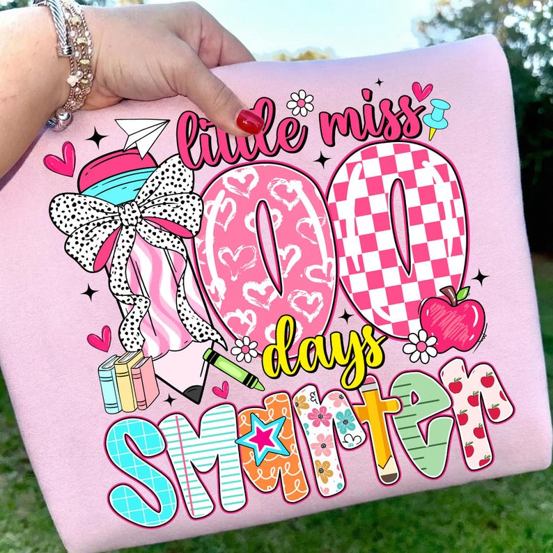 May include: Pink graphic t-shirt with the text "Little Miss 100 Days Smarter." The design includes hearts, a bow, books, an apple, and a crayon. The shirt is a light pink color.