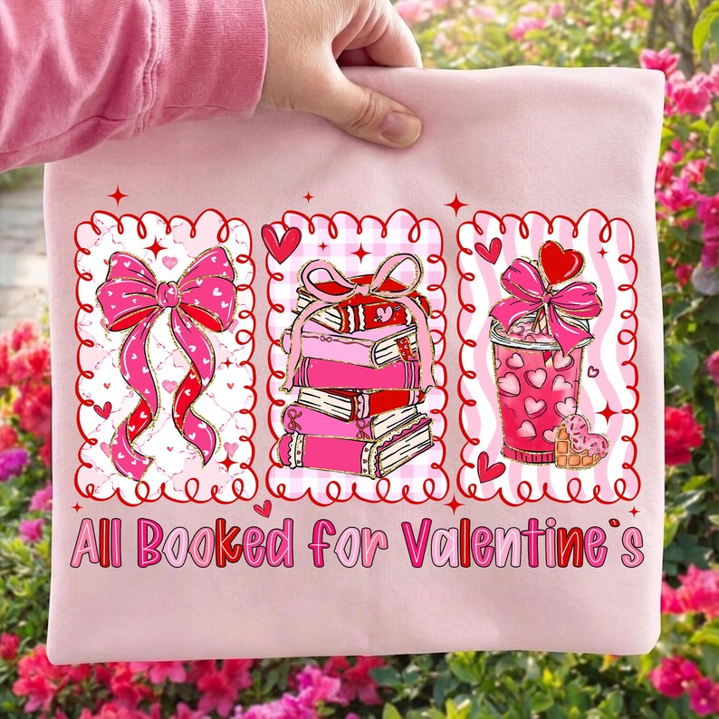 May include: A light pink sweatshirt featuring three Valentine's Day themed graphics. The graphics include a bow, a stack of books, and a drink, all in shades of pink and red. The text "All Booked for Valentine's" is printed at the bottom.