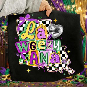 May include: Black tote bag with the text "La Weezy Ana" in colorful, block letters. The design includes a disco ball heart, a crown, and a checkered pattern, with Mardi Gras colors of purple, green, and gold.