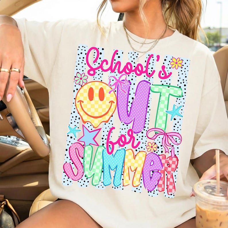 Puede incluir: Camiseta color crema con un gr&aacute;fico colorido que dice "School's Out for Summer". El dise&ntilde;o incluye una carita sonriente, estrellas, lazos y flores. El texto y los gr&aacute;ficos son en rosa brillante, morado, verde y amarillo.