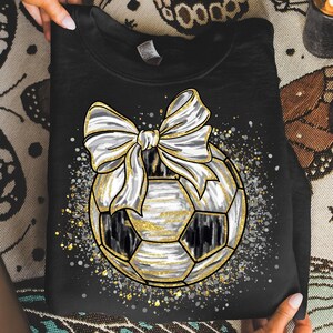 K&ouml;nnte beinhalten: Schwarzes Sweatshirt mit einem Fu&szlig;ball-Design, einer wei&szlig;en Schleife und goldenen Akzenten. Der Fu&szlig;ball hat schwarz-wei&szlig;e Paneele mit goldenen Details. Das Design ist von einem gesprenkelten Muster aus Gold und Silber umgeben.