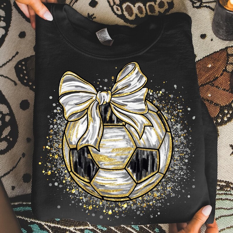 K&ouml;nnte beinhalten: Schwarzes Sweatshirt mit einem Fu&szlig;ball-Design, einer wei&szlig;en Schleife und goldenen Akzenten. Der Fu&szlig;ball hat schwarz-wei&szlig;e Paneele mit goldenen Details. Das Design ist von einem gesprenkelten Muster aus Gold und Silber umgeben.