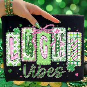 Op de afbeelding: Zwart T-shirt met de woorden "LUCKY VIBES" in een groen en roze ontwerp. De letters zijn versierd met klavers en patronen. Boven de letter "G" staat een roze strik. Het shirt is versierd met klavers.