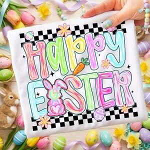 Może przedstawiać: Biała koszulka z kolorową grafiką "Happy Easter". Projekt zawiera pastelowe litery, marchewkę, pisanki i kr&oacute;lika. Koszulka ma czarno-białą szachownicę i jest otoczona dekoracjami wielkanocnymi.