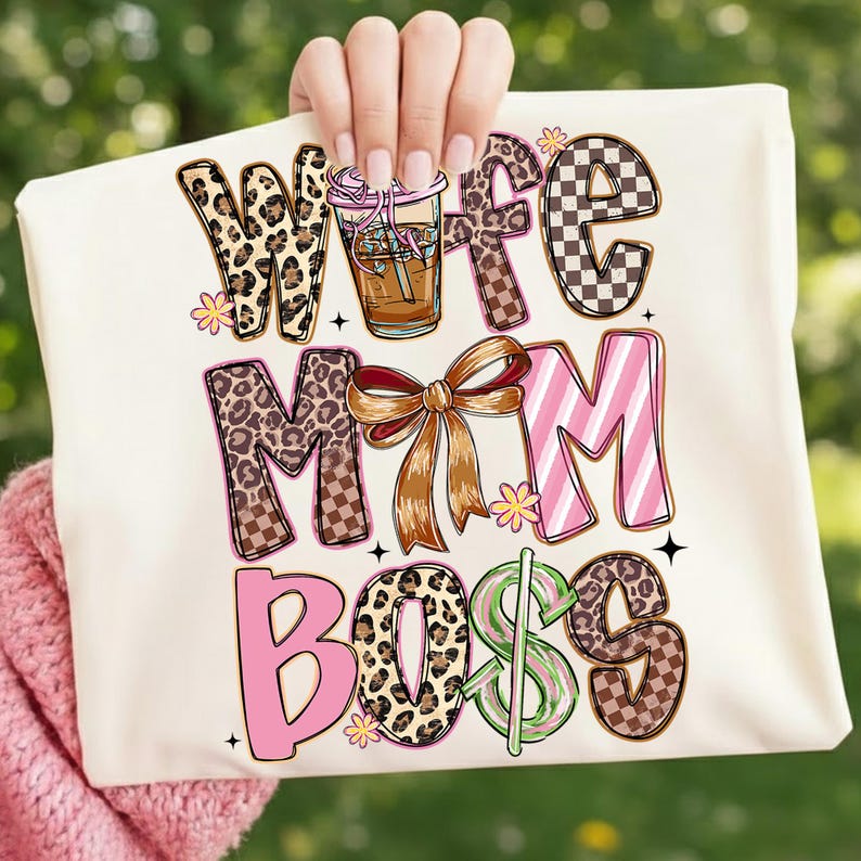 May include: Cream-colored tote bag with the words "Wife Mom Boss" in various decorative fonts. The letters feature leopard print, checkered patterns, and pink stripes. A coffee drink and a bow are also included in the design.