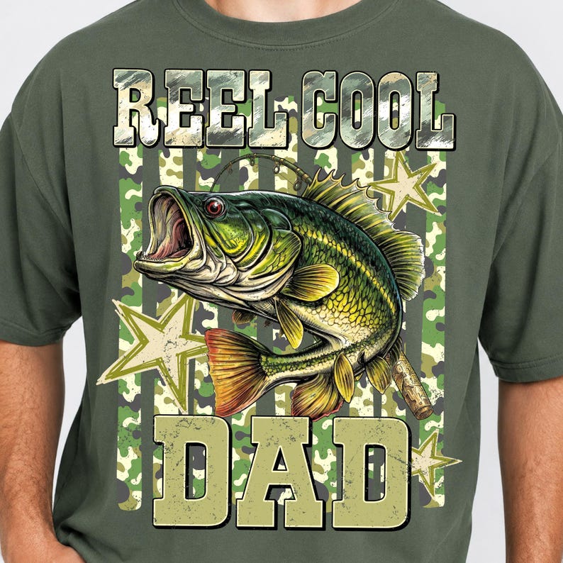 May include: Olive green t-shirt with a graphic of a largemouth bass and the words "REEL COOL DAD" in a camouflage design. The fish is green and yellow with an open mouth. Stars are in the background.