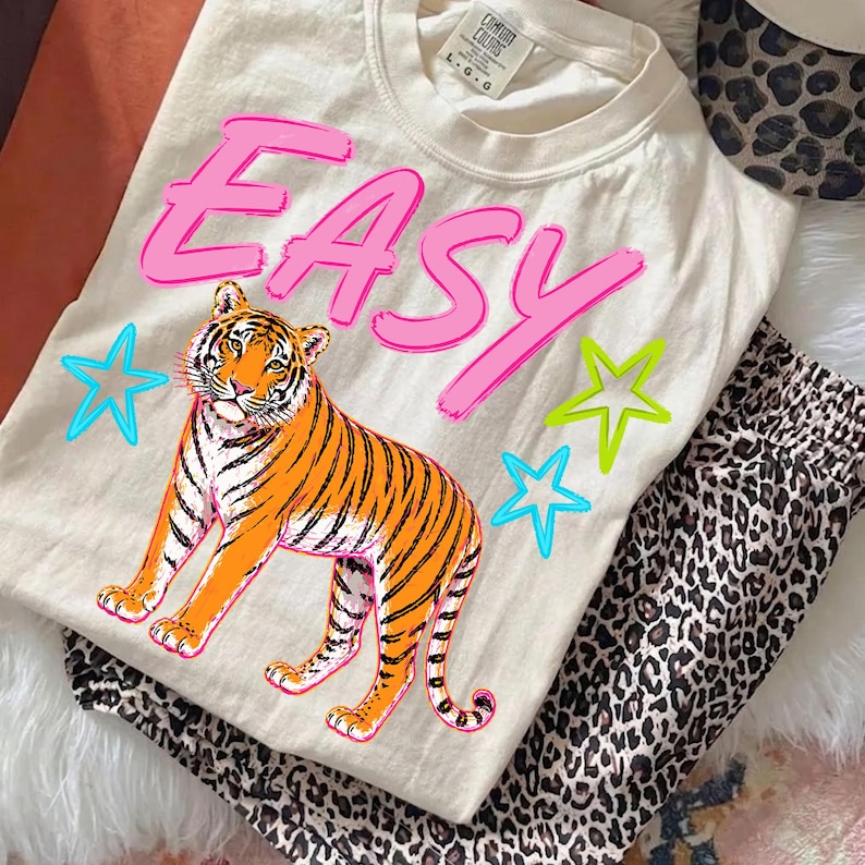 May include: Cream-colored t-shirt with the word "EASY" in pink, a tiger graphic, and star accents. Paired with leopard print pants and a leopard print accessory.