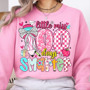 May include: Pink sweatshirt with a colorful graphic celebrating 100 days of school. The design includes the words "Little Miss 100 Days Smarter" with hearts, flowers, and an apple. A large bow and books are also featured.